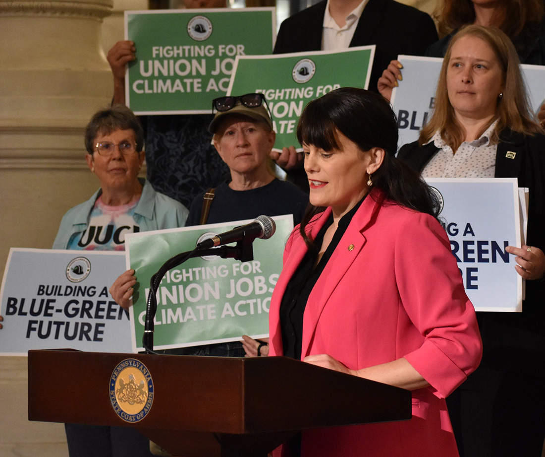 A woman speaks at a podium in front of a group of people holding signs.