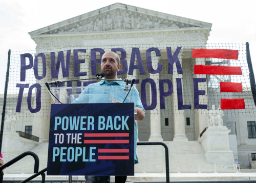 A man stands at a podium in front of the Supreme Court with signs that read "Power back to the people."