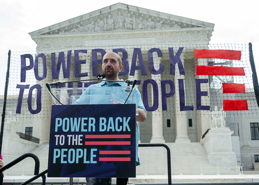 A man stands at a podium in front of the Supreme Court with signs that read "Power back to the people."