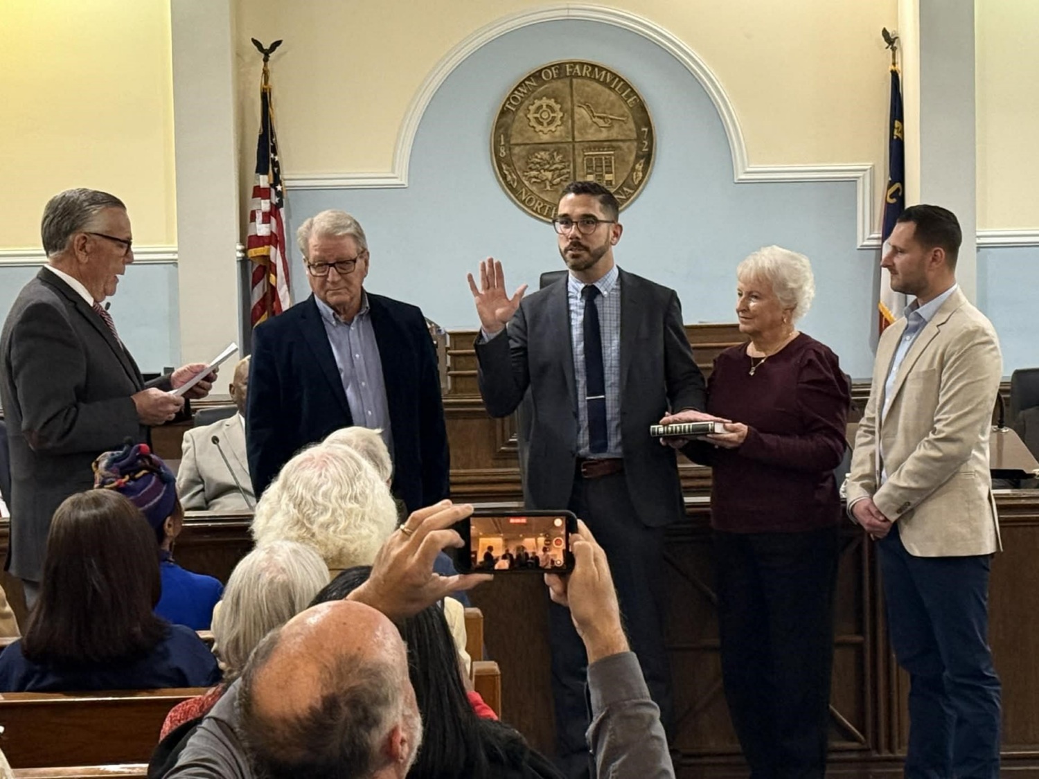 Standing in town council chambers, a man with his right hand raised takes the oath of office while supporters look on.