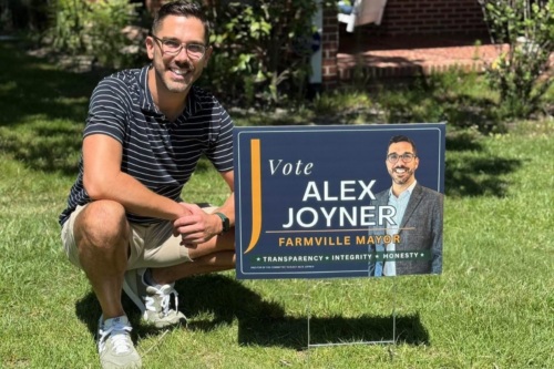 A man poses smiling on a green lawn next to a campaign yard sign bearing his portrait and reading "Vote Alex Joyner, Farmville Mayor, Transparency, Integrity, Honesty"
