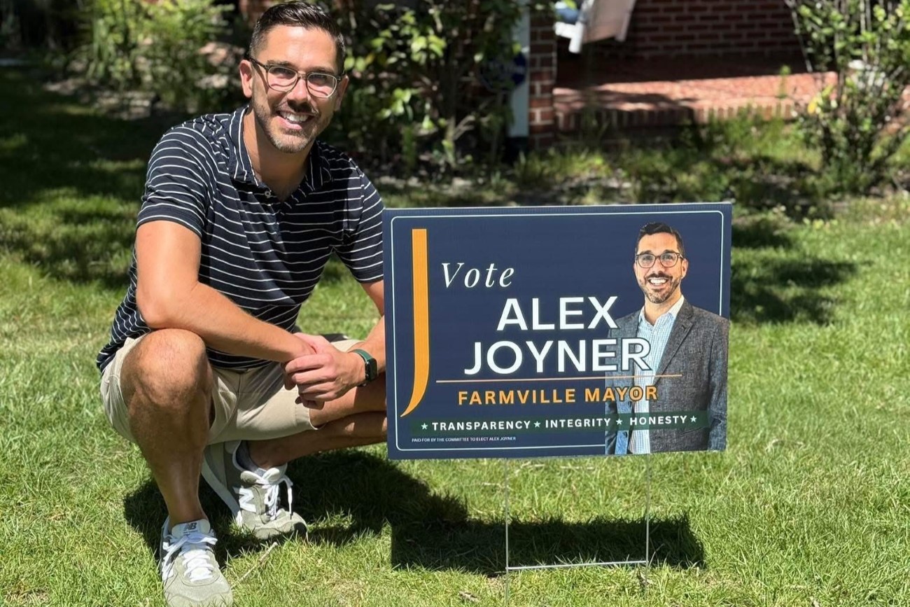 A man poses smiling on a green lawn next to a campaign yard sign bearing his portrait and reading "Vote Alex Joyner, Farmville Mayor, Transparency, Integrity, Honesty"