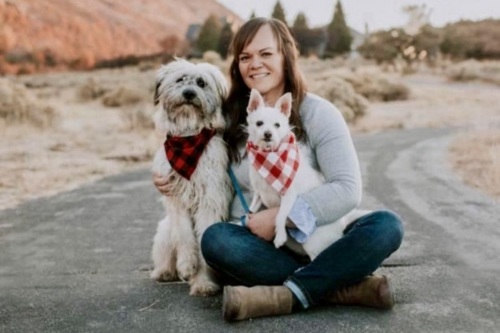 On a park path, a smiling woman sits with two white dogs sporting red checkered bandanas.