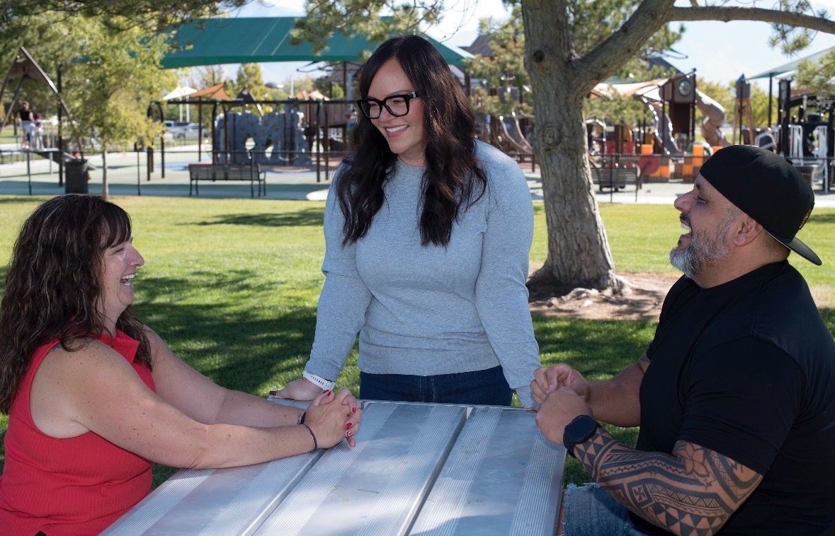 In a town park with a playground in the background. a smiling woman stands next to a man and a woman seated at a picnic table, both laughing.