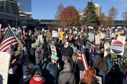 A crowd bundled in jackets and carrying American flags and protest signs at an autumn rally in downtown Anchorage.