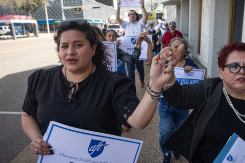 Two women lead a group marching down a town street, holding signs reading "Protect Our Kids."