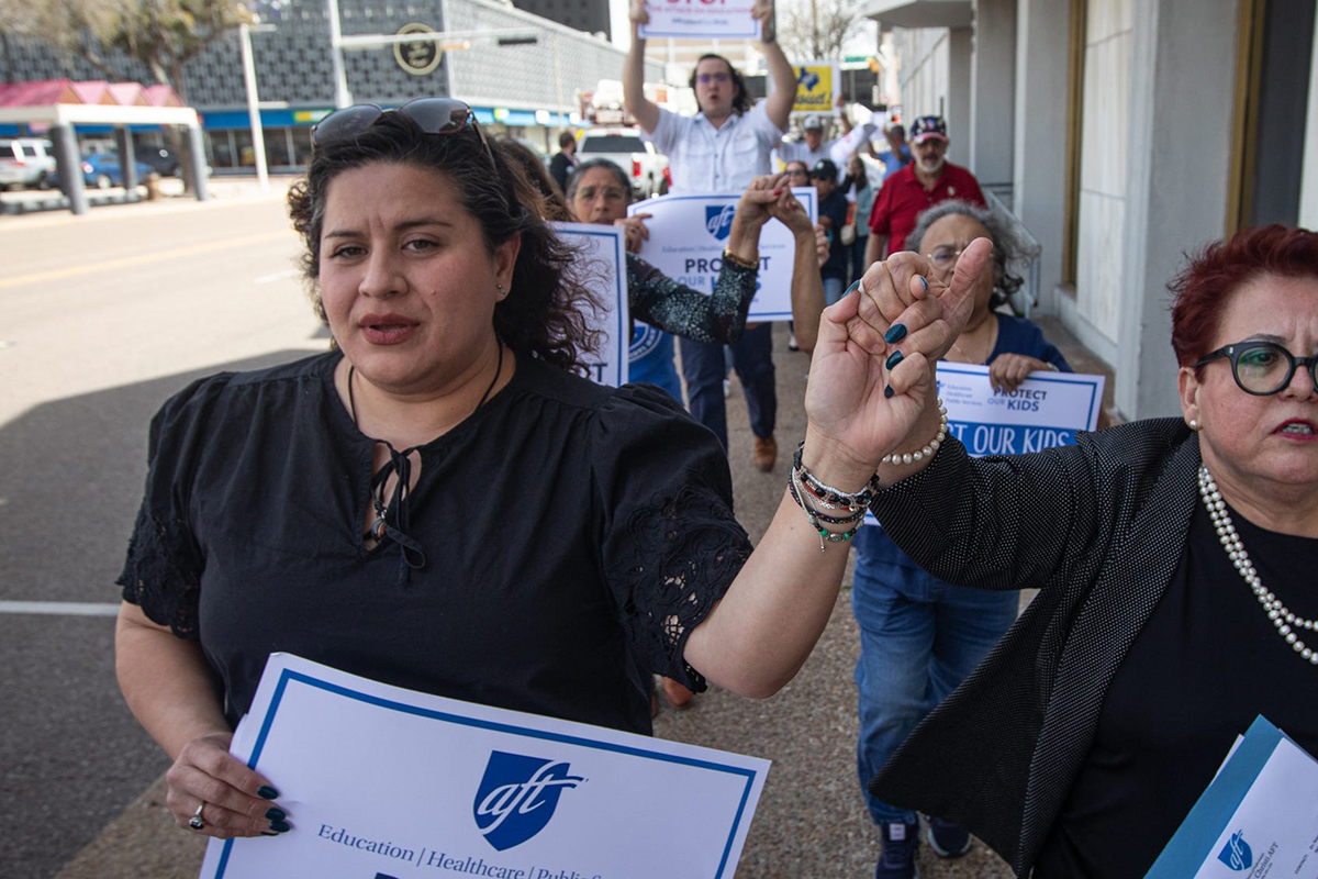 Two women lead a group marching down a town street, holding signs reading "Protect Our Kids."