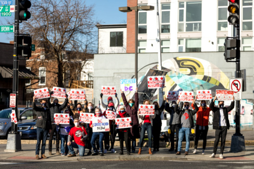 A group demonstrates on a DC street corner, holding white signs with red stars and stripes in the style of the DC flag. The signs read "Statehood for the People of DC."