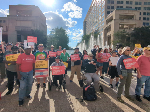 A group of advocates from Indiana Conservation Voters gather outside to protest redistricting in front of a fountain and buildings.