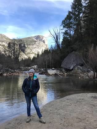 A woman stands in front of a body of water and trees.