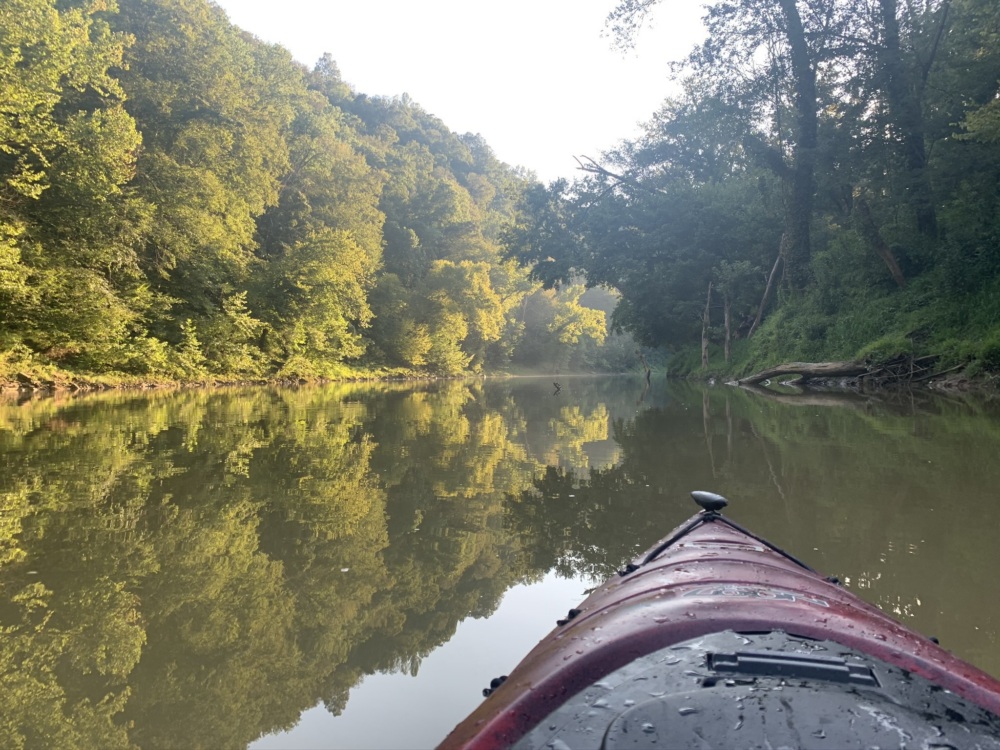 A tree lined body of water with the front of a kayak in the foreground.