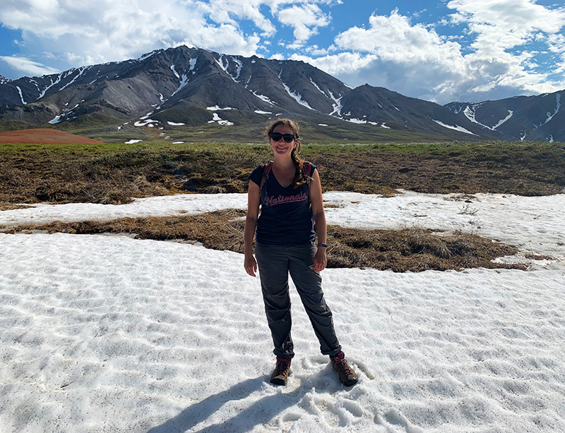 A woman stands on snow in front of a mountain.