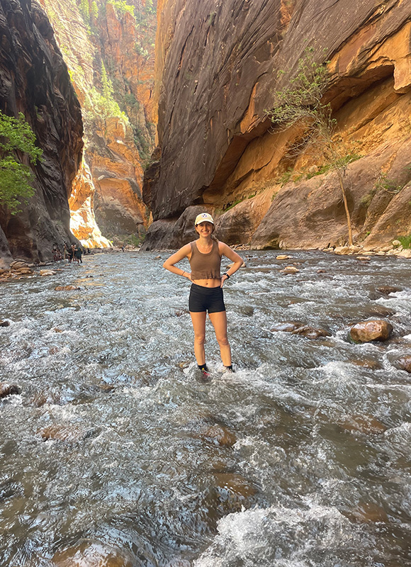 A woman stands in a shallow river between 2 rock formations.