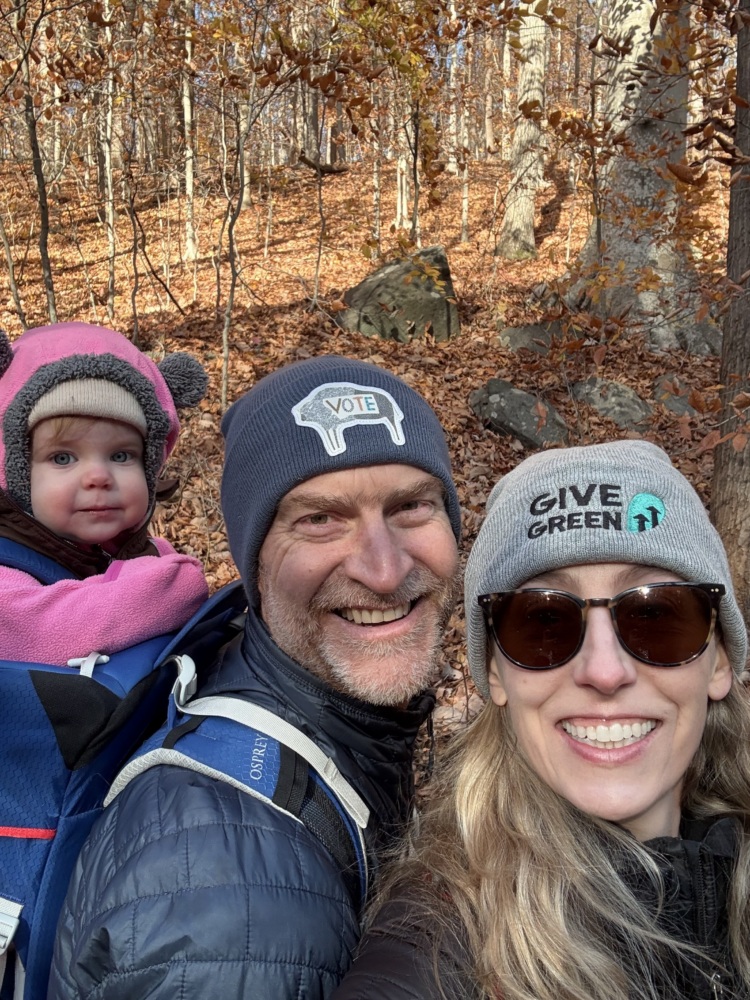A man, a woman, and a baby smile in the woods.