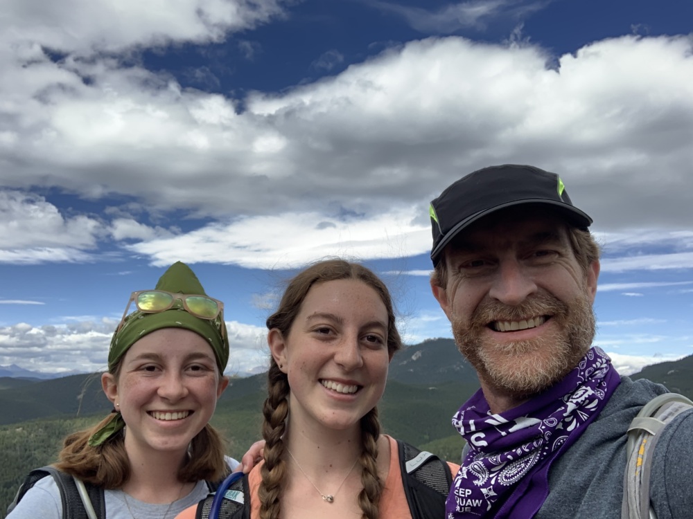 Two young women and a man smile in front of a mountain backdrop.