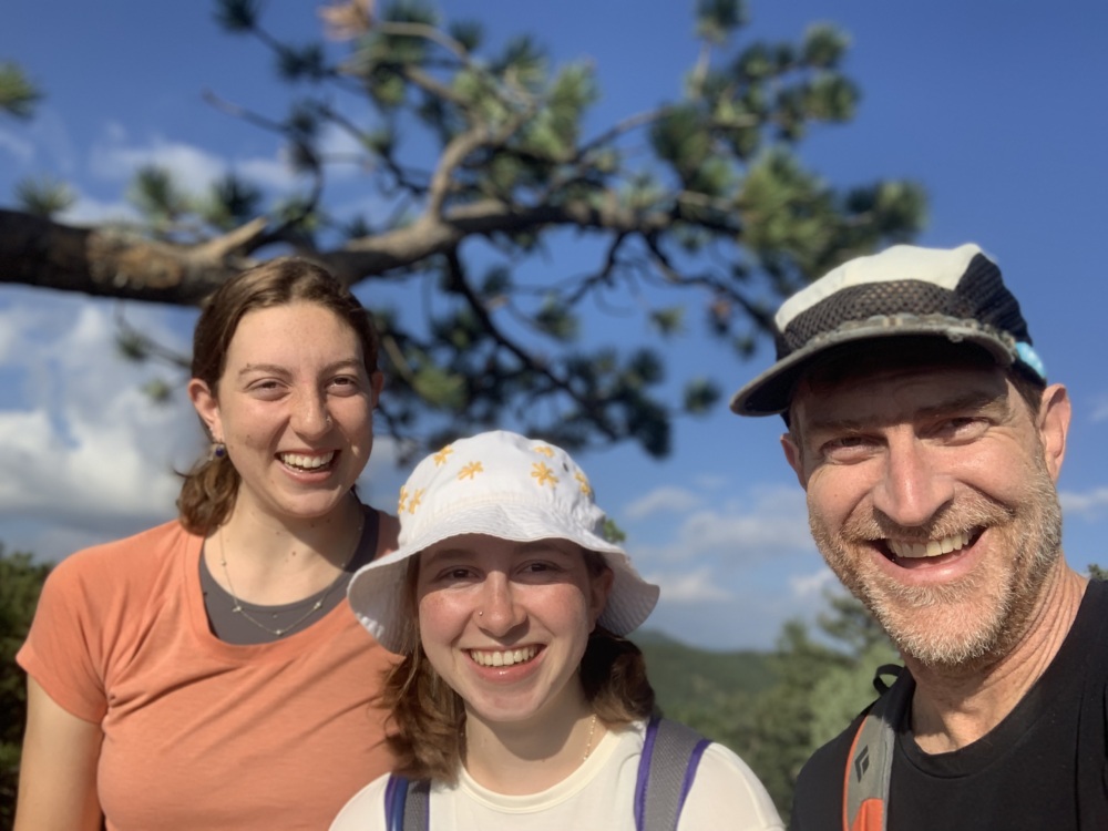 Two young women and a man smile in front of a green landscape.