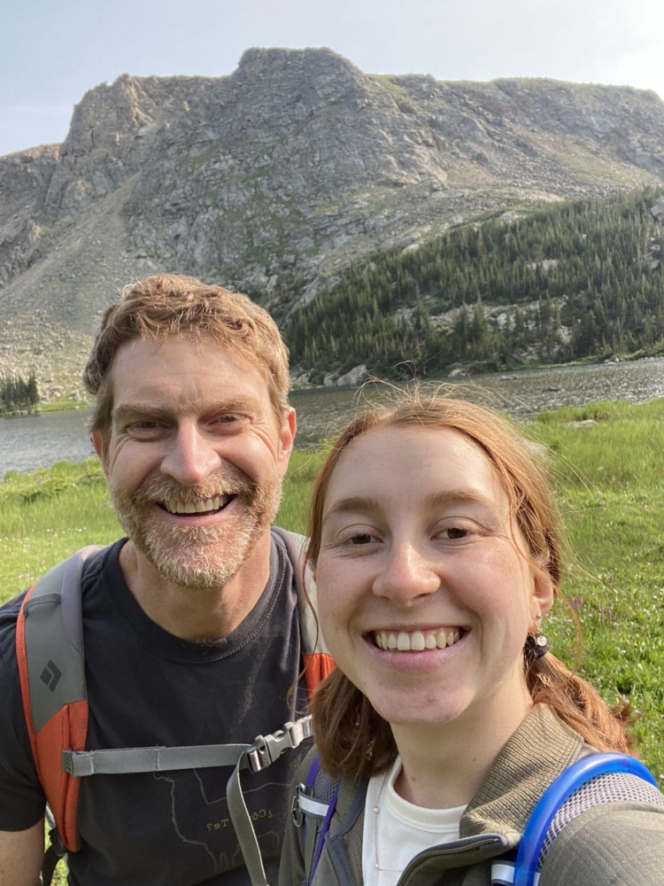 A man and young woman smile in front of a green mountain backdrop.